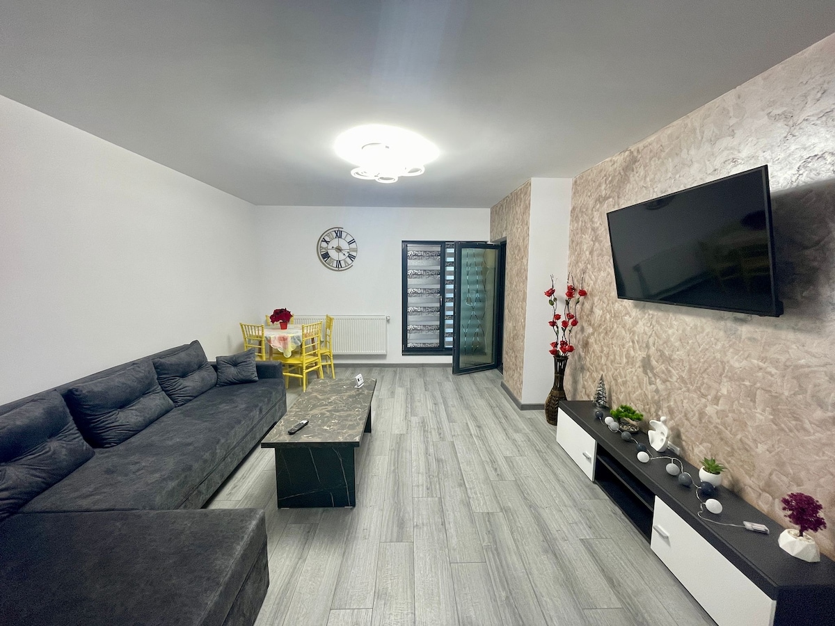 A spacious living area features a large, dark sectional sofa alongside a sleek coffee table. A modern television is mounted on the wall, and a dining area with yellow chairs is visible. Decorative plants and a wall clock add subtle touches to the ambiance.
