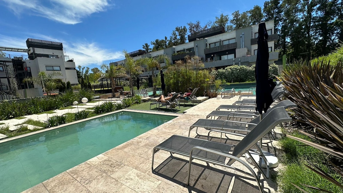 A serene outdoor pool area is framed by well-maintained gardens and modern buildings. Lounge chairs are positioned along the pool's edge, providing a comfortable spot for relaxation. A few umbrellas offer shade, while the clear blue sky adds to the tranquil setting.