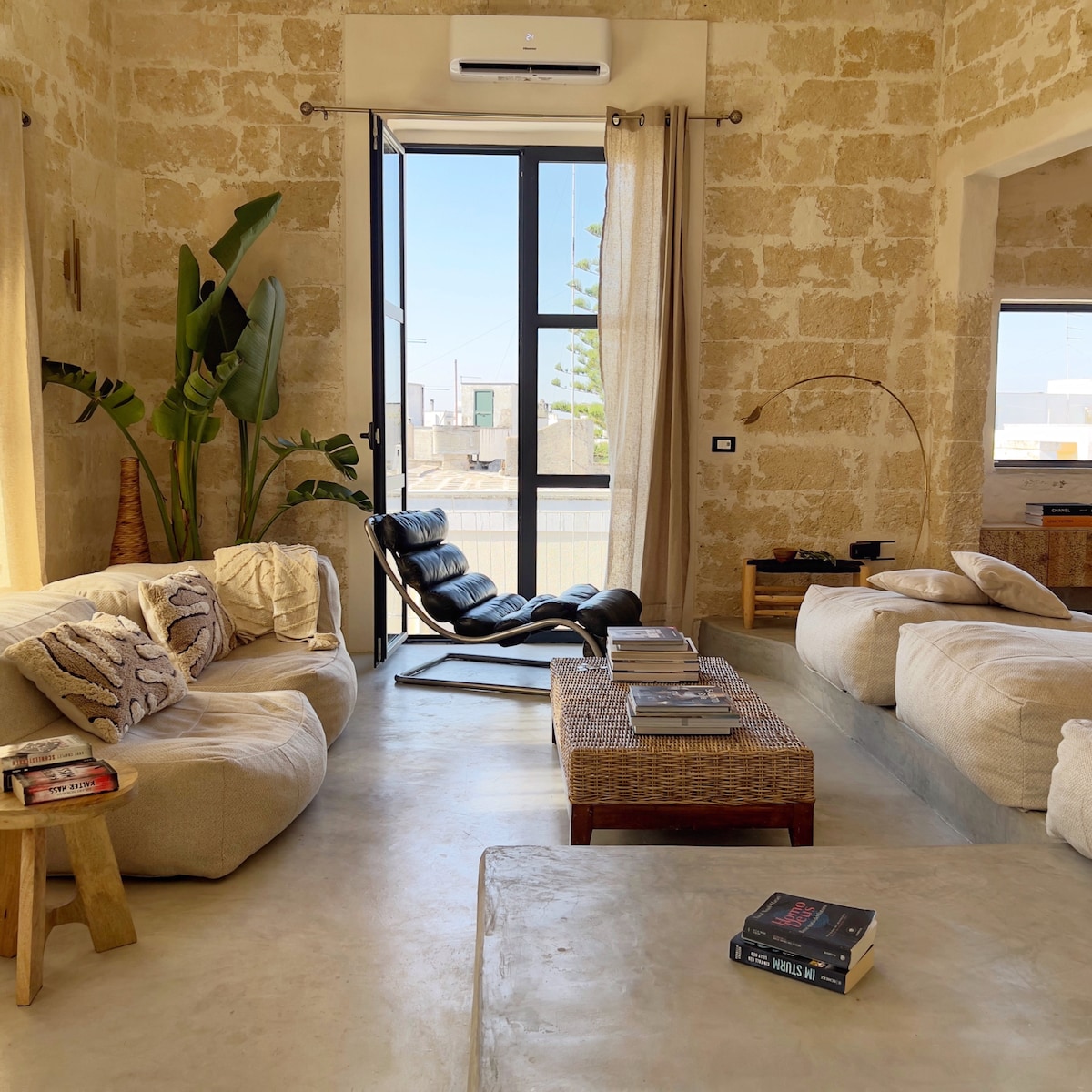 A spacious living area features a mix of comfortable seating options, including a casual sofa and a sleek black lounge chair. Large windows allow natural light to fill the space, highlighting the textured stone wall and earthy tones. A coffee table is arranged with books and decorative items.