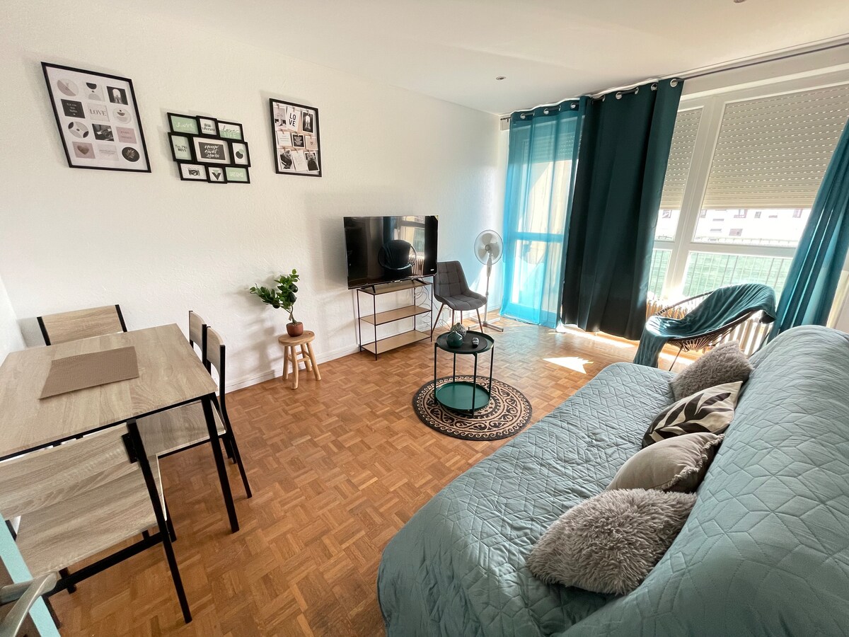 A living space is showcased with a comfortable sofa and a modern TV stand featuring a flat-screen television. A small dining table is positioned next to the seating area. Natural light fills the room through large windows, complemented by teal curtains and a patterned rug on the wooden floor.