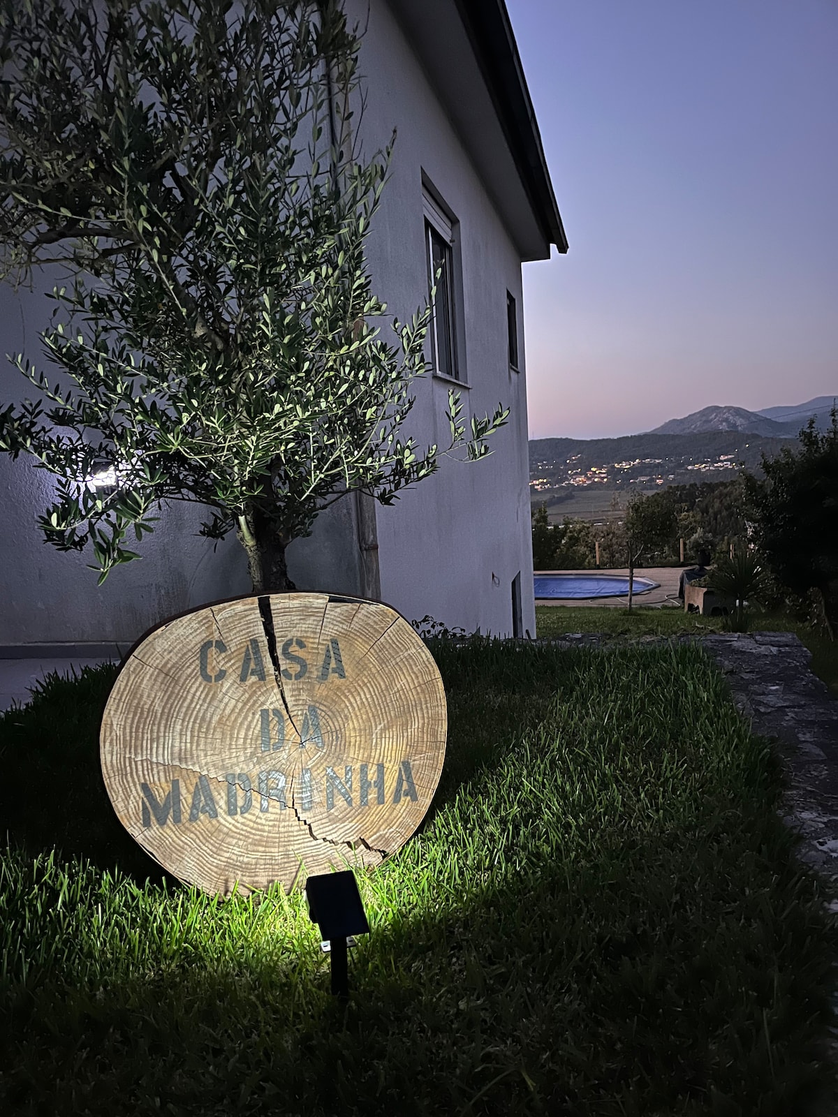 A wooden sign displaying the name 'Casa da Madrinha' is illuminated softly at night, positioned on the lawn next to an olive tree. The villa's exterior is visible in the background, with distant hills and the outline of a pool under the evening sky.