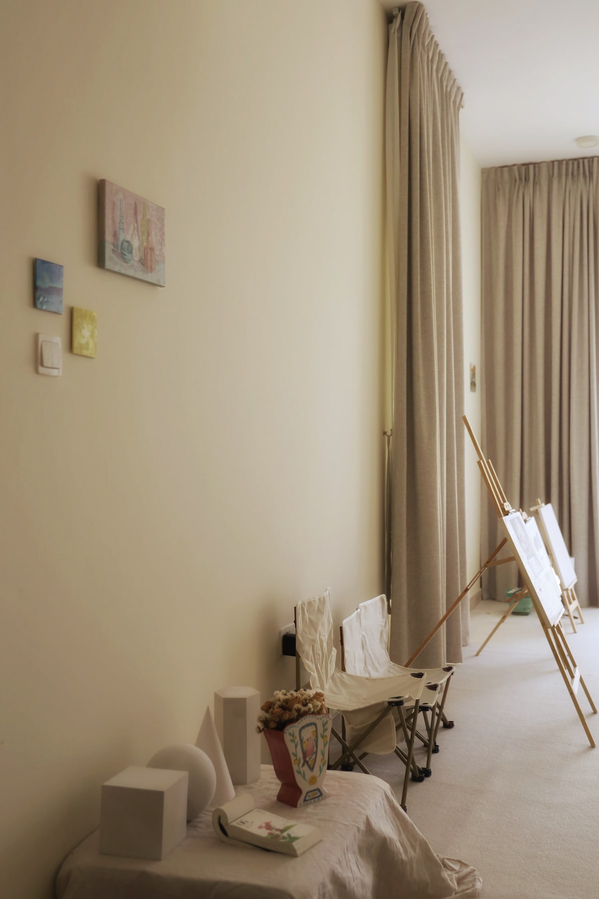A soft-toned wall showcases small art pieces that add character to the space. Nearby, two lightweight folding chairs are positioned beside a table. Natural light streams in through tall curtains, illuminating an easel with blank canvases, suggesting a creative and calm environment.