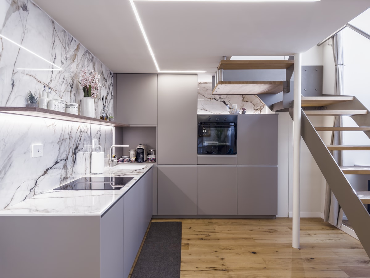 The sleek kitchen features modern cabinetry in muted tones, accented by a striking marble backsplash. High-tech appliances, including a built-in oven, are visible. Warm wooden flooring adds a natural element to the space, with soft lighting enhancing the contemporary design.