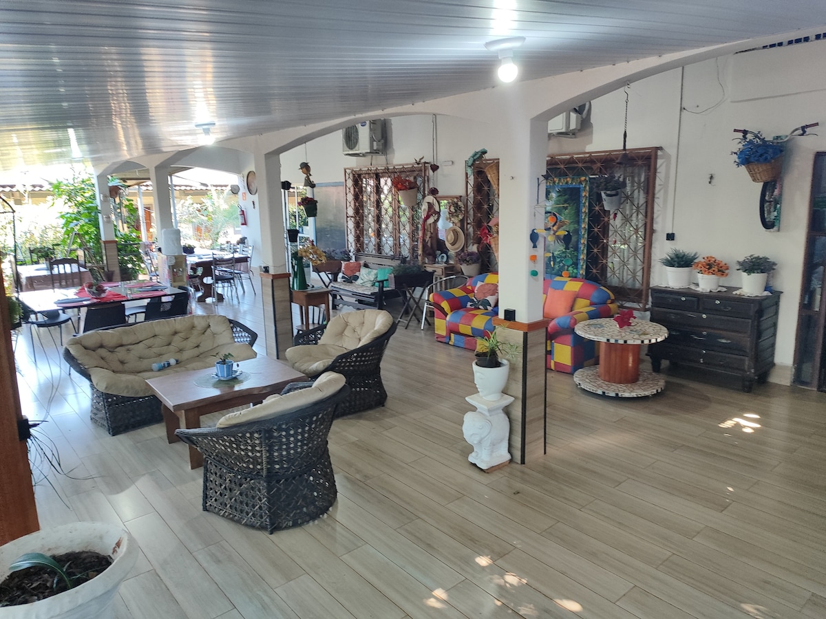 An open-concept living area features a variety of seating options, including wicker chairs and a colorful sofa. Natural light filters through large windows, illuminating wooden furniture and decorative elements. Greenery is visible throughout, adding a touch of nature to the inviting space.