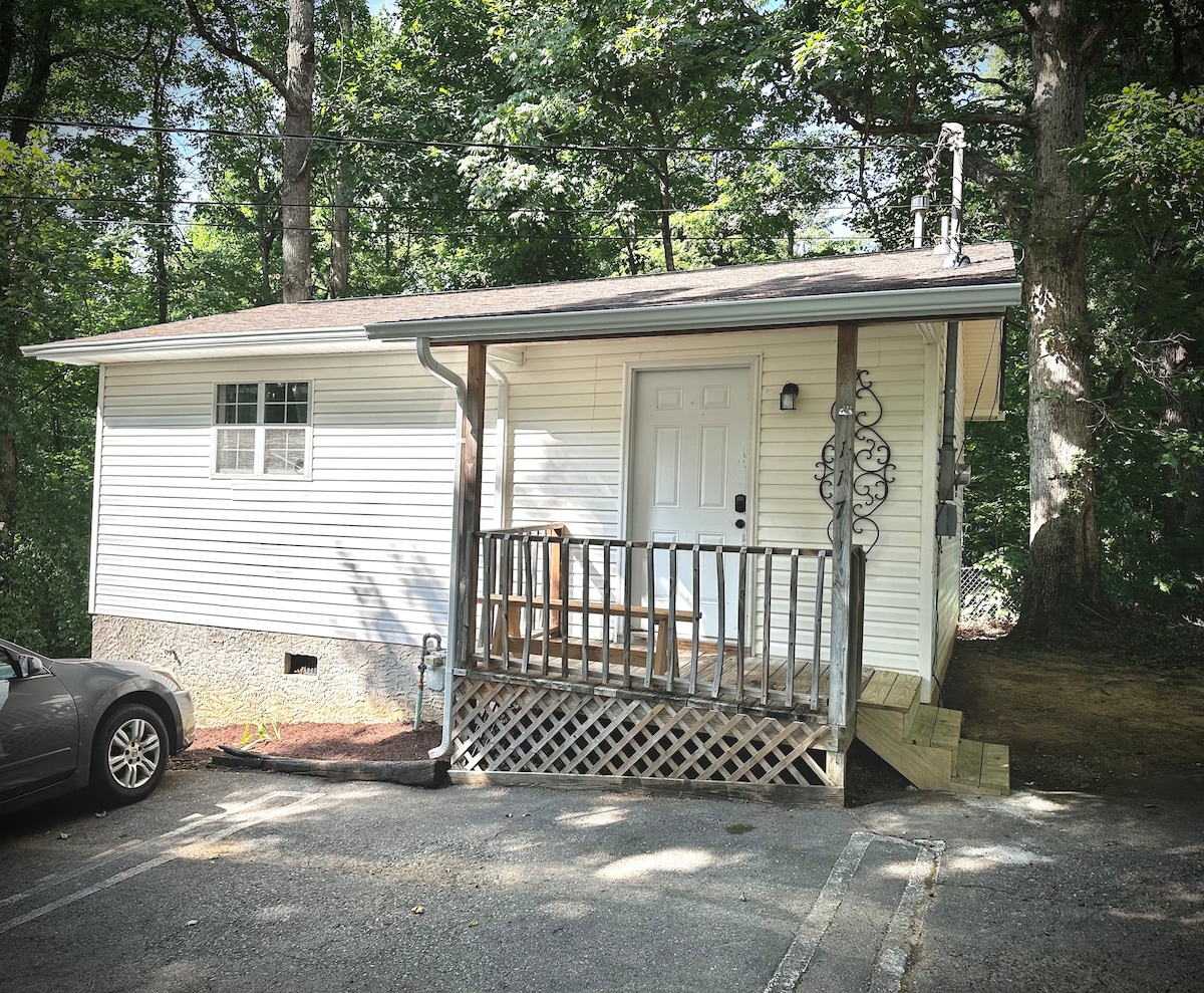 The exterior of the unit is shown, featuring a well-maintained white facade with a welcoming front porch. A driveway is visible, accommodating parking nearby. Lush green trees surround the property, providing a sense of privacy and tranquility.