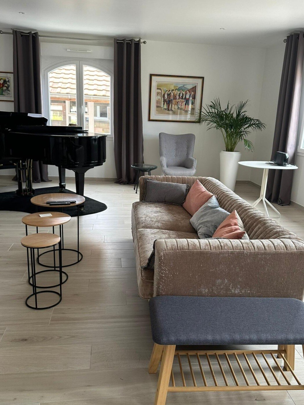 A well-appointed living area features a plush sofa with accent pillows, complemented by two stylish side tables. A light-colored armchair and a small round table are positioned nearby. A grand piano stands elegantly in one corner, adding a touch of sophistication.