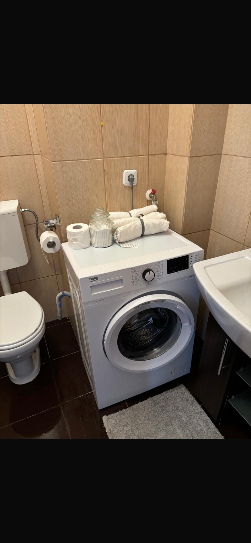 A compact laundry area is displayed, featuring a white washing machine positioned next to a modern sink. Neatly folded towels and a roll of toilet paper are arranged on top. The walls are covered in light-colored tiles, complemented by a dark floor.