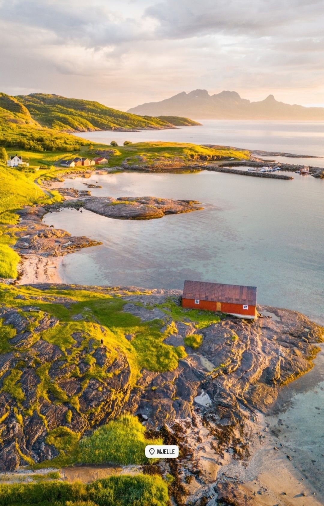An aerial view captures a charming red house nestled on rocky terrain near the shoreline. Surrounding the house are lush green areas and gently sloping hills leading to a serene beach and calm waters, with distant mountains rising in the background under a soft, cloudy sky.