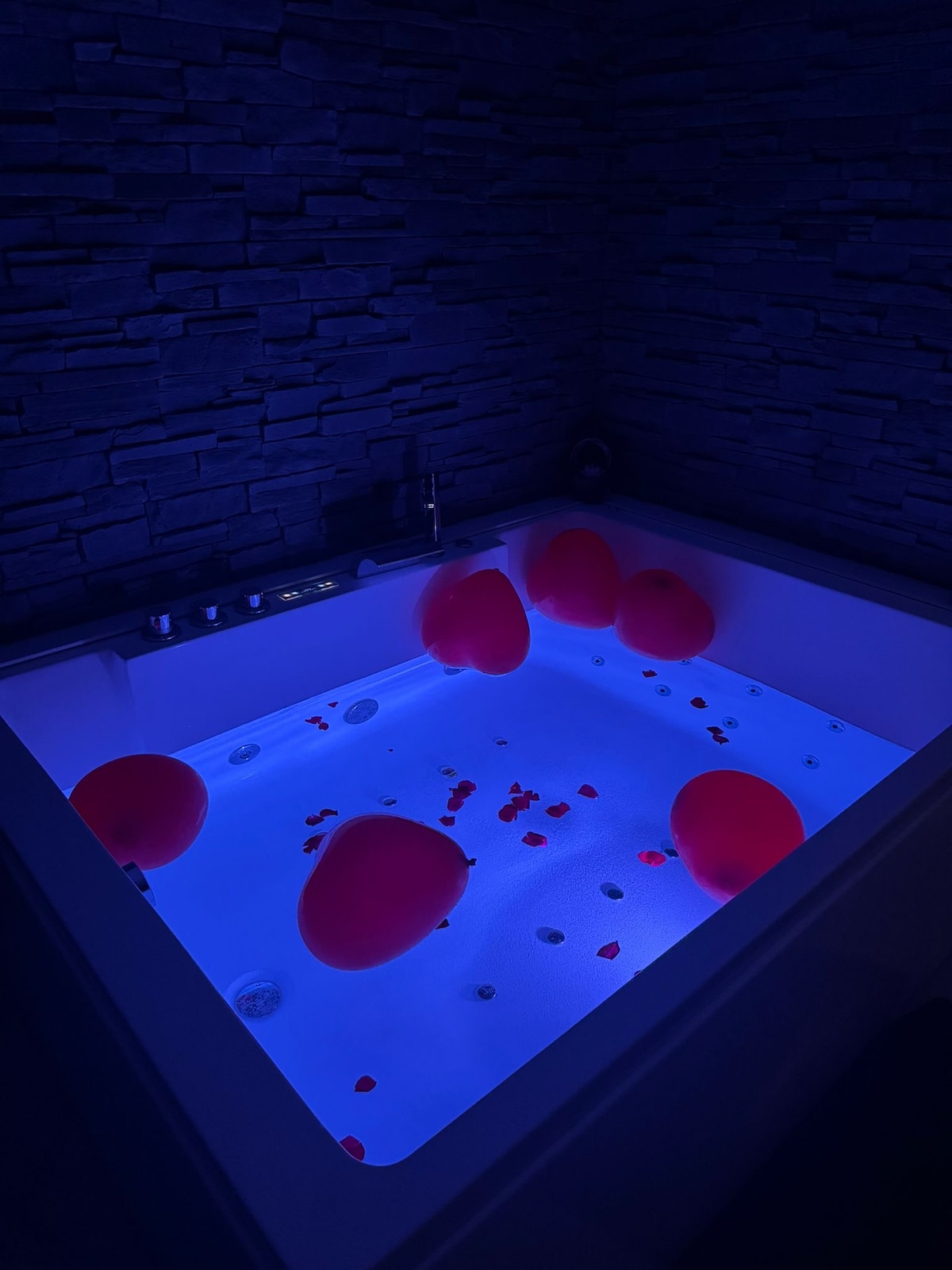 A modern jacuzzi is illuminated with soft blue lighting, enhancing the serene ambiance. The surface is decorated with red balloons and rose petals, creating a festive and inviting scene. The textured stone wall provides a stylish backdrop, contributing to the overall aesthetic.