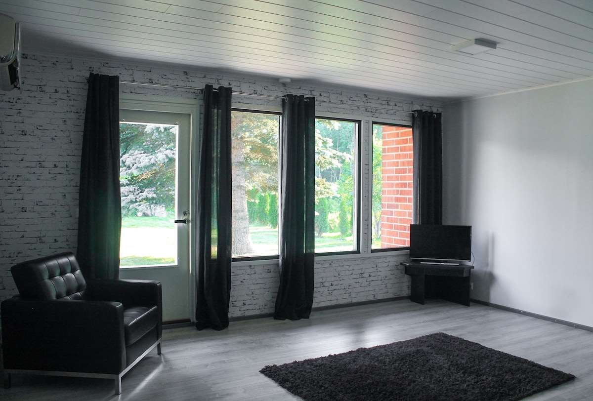 A spacious living area features large windows that allow natural light to fill the room. Dark curtains frame the windows, and a black armchair sits in one corner atop a soft area rug. A television is positioned near the window, providing entertainment options.