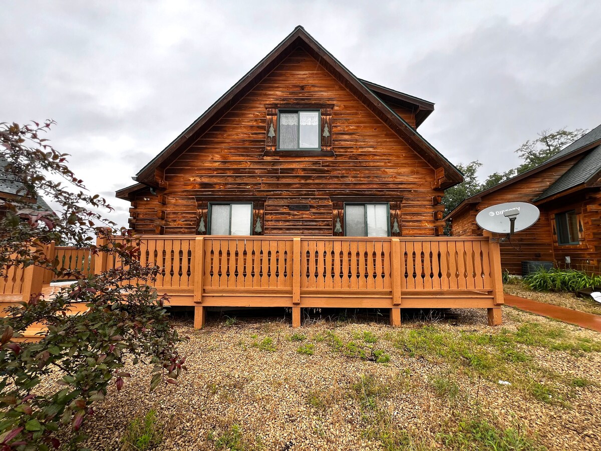 A wooden cabin exterior is showcased, featuring a warm, rustic finish. The structure has a sloped roof and multiple windows, allowing natural light to enter. A spacious wooden deck runs along the front, bordered by a low fence, providing a welcoming outdoor area.