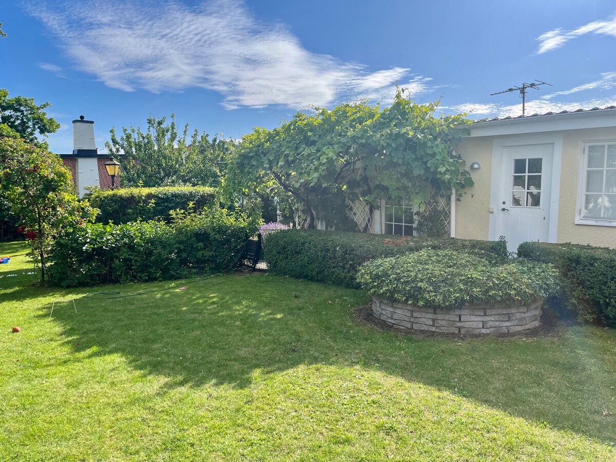 A well-maintained garden surrounds a charming guesthouse, featuring hedges and a stone planter. Lush green grass covers the area, while a trellis supports vibrant vines. Sunlight filters through the leaves, creating a serene outdoor space ideal for relaxation.