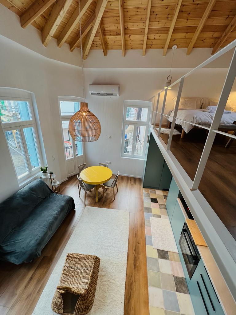 An airy living space features high, wooden ceilings and large windows allowing natural light to fill the room. A cozy sofa is complemented by a round yellow dining table, with a woven basket accentuating the floor design. The upper level houses a sleeping area, visible from below.