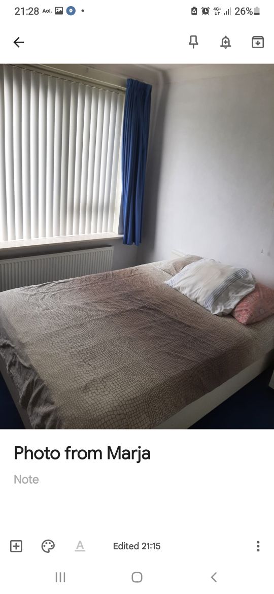 A serene sleeping area features a spacious bed adorned with neutral-toned bedding and multiple pillows. Natural light filters through vertical blinds, highlighting the soft textures of the bedspread. The room is complemented by light-colored walls and a blue curtain, creating a calming environment.