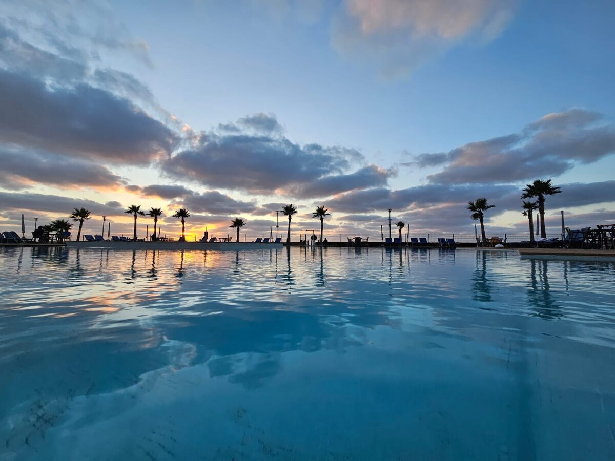 A tranquil pool reflects the soft hues of sunset, surrounded by palm trees that add to the serene setting. The sky is adorned with clouds, enhancing the peaceful atmosphere of the outdoor space. Lounge chairs are visible in the background, suggesting relaxation.