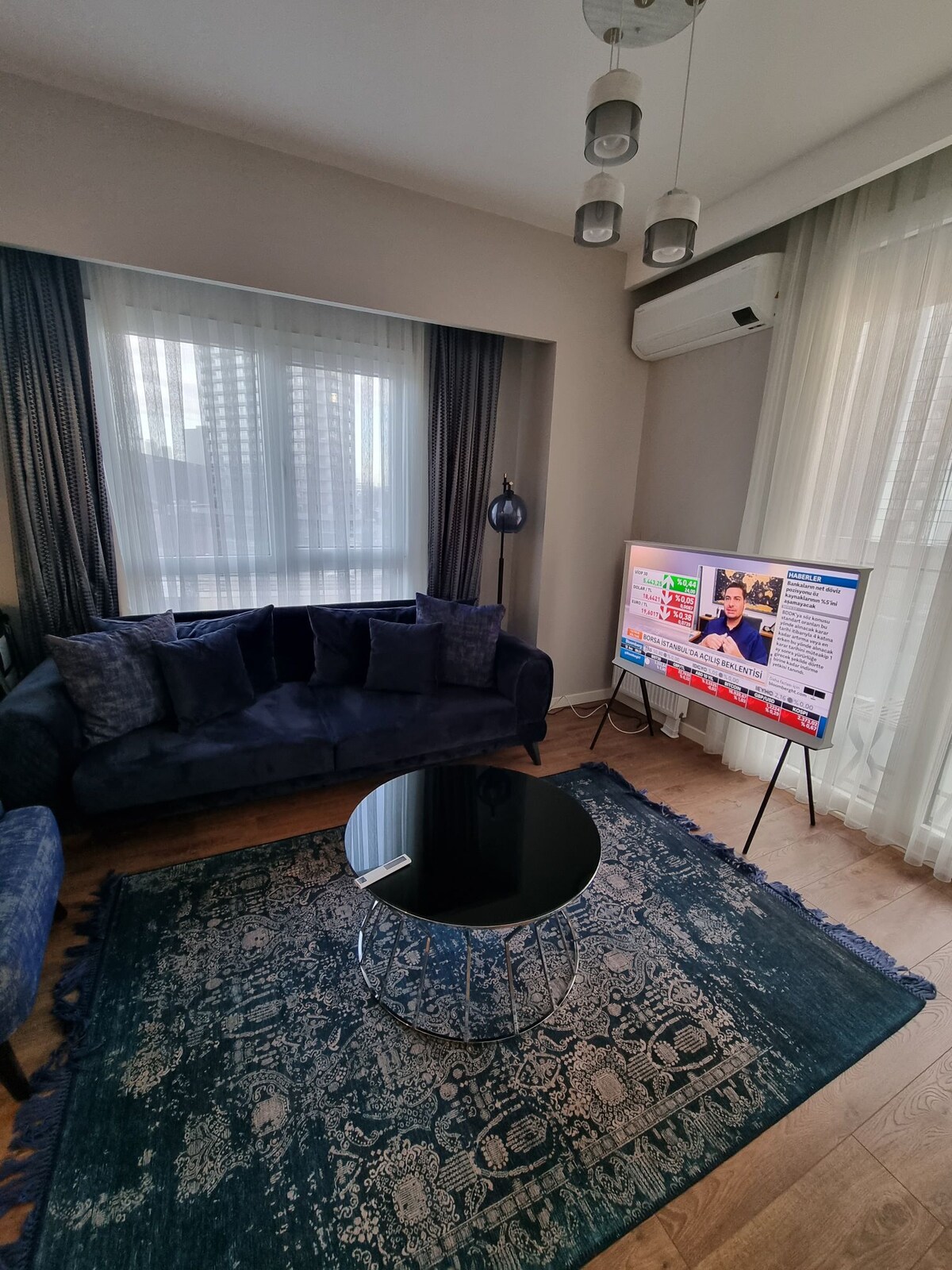 A modern living area features a plush dark blue sofa paired with a decorative armchair. A glass coffee table sits on a traditional patterned rug. A large television displays vibrant graphics, while natural light filters through sheer curtains, creating a bright and inviting space.