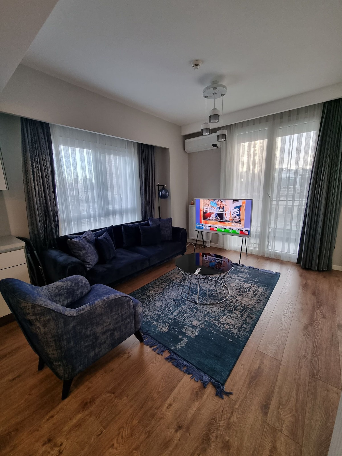 A living area features a dark blue sofa and an armchair, complemented by a circular glass coffee table. A TV stands on a stand, showing colorful content. Two large windows with sheer curtains allow natural light to fill the space, highlighting wooden flooring.