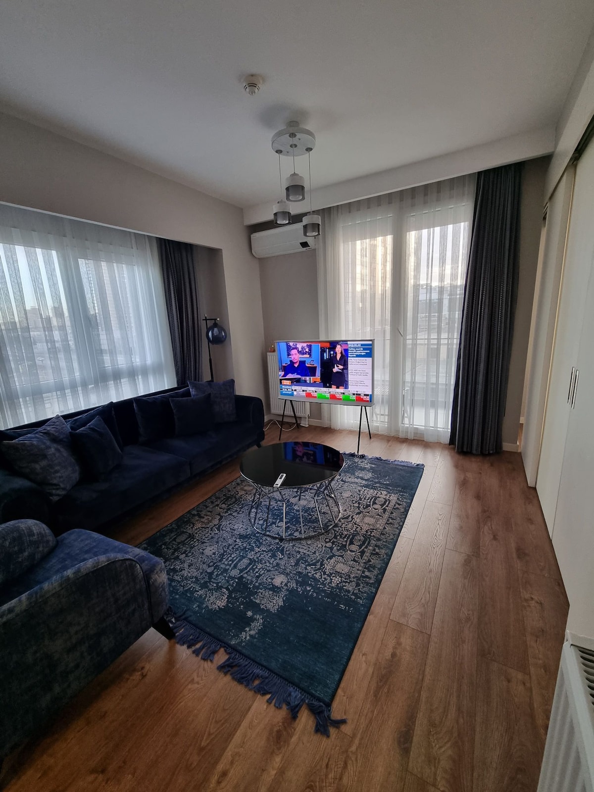A modern living area features a dark blue sectional sofa arranged around a glass coffee table. A colorful television display is visible, and large windows dressed with sheer curtains allow natural light to fill the space. A soft area rug adds texture to the wooden floor.