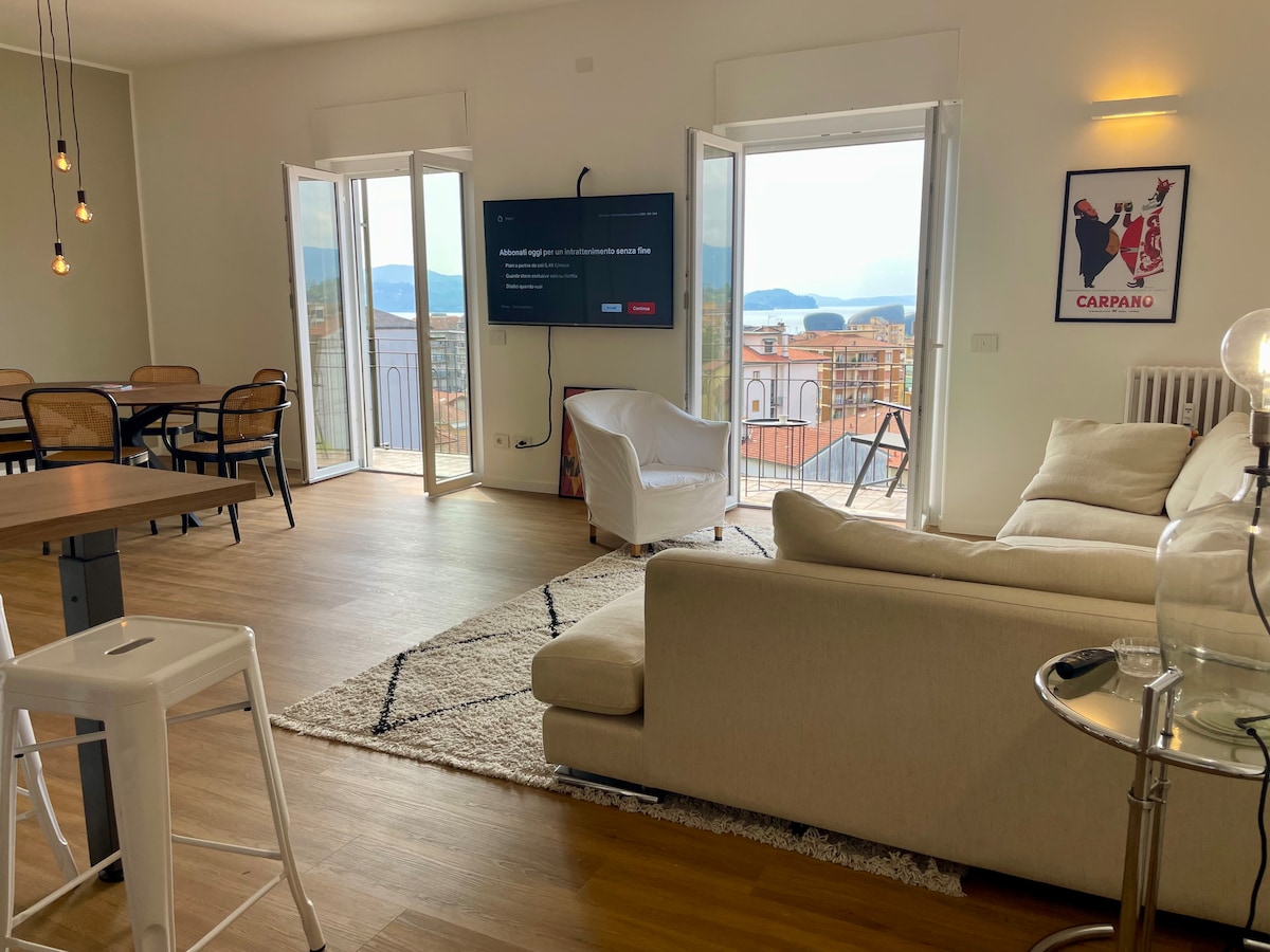 A bright living area features a spacious sofa, a television mounted on the wall, and three large windows that open to a panoramic lake view. A dining table is positioned nearby, with several chairs arranged around it. Natural light floods the room, creating an airy atmosphere.