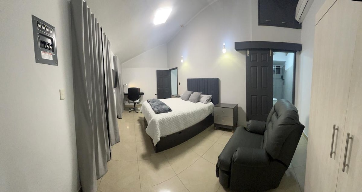 A spacious bedroom features a comfortable bed with light bedding, accompanied by a dark gray armchair and a small nightstand. Soft gray curtains frame the area, and a desk is positioned nearby, creating a functional workspace. Subtle lighting enhances the neutral color palette.