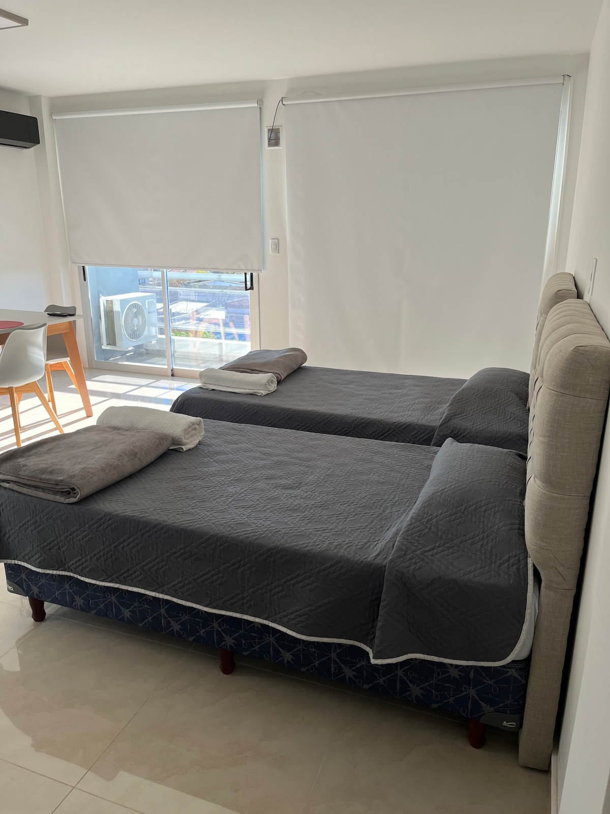 A comfortable sleeping area features two individual beds dressed in dark quilted bedding. Soft towels are neatly arranged on each bed. A bright window allows natural light to fill the space, while neutral-colored walls contribute to a serene environment.