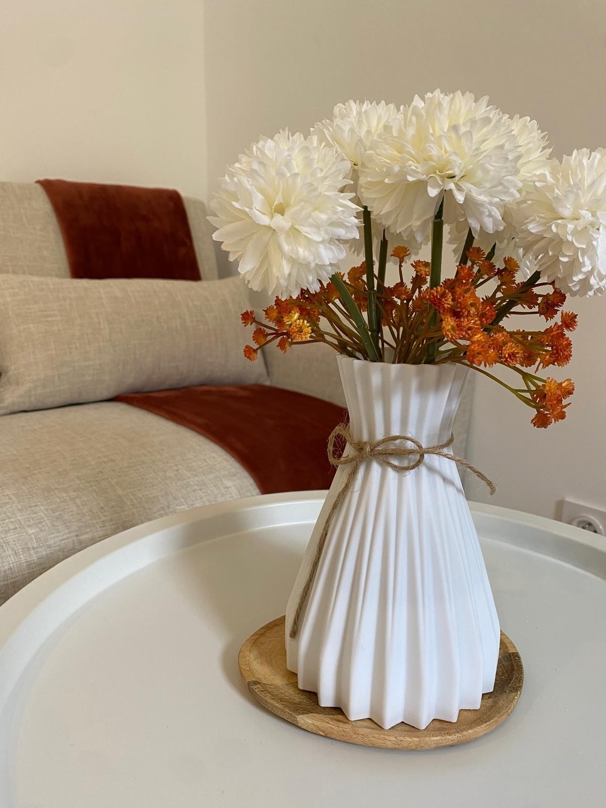 A white vase featuring pleated design holds an arrangement of white flowers and orange accents. The vase is positioned on a circular wooden tray atop a white table, with a beige and rust-colored couch visible in the background.