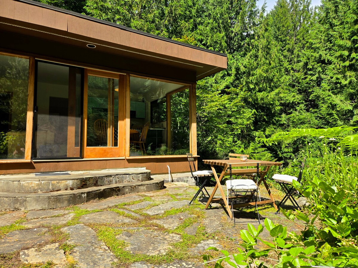 An outdoor seating area is visible, featuring a small wooden table and four chairs positioned on a stone patio. Lush greenery and trees frame the cabin, enhancing the natural setting. Large glass doors invite the outside in, while a few steps lead to the entrance.