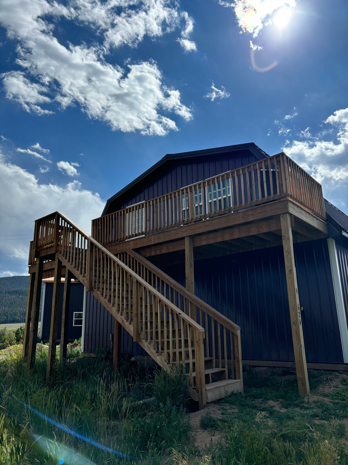 The exterior of the loft features a wooden staircase leading up to a balcony, surrounded by lush greenery and under a clear blue sky. The building's dark exterior contrasts with the natural surroundings, while the sun casts a bright light over the structure.