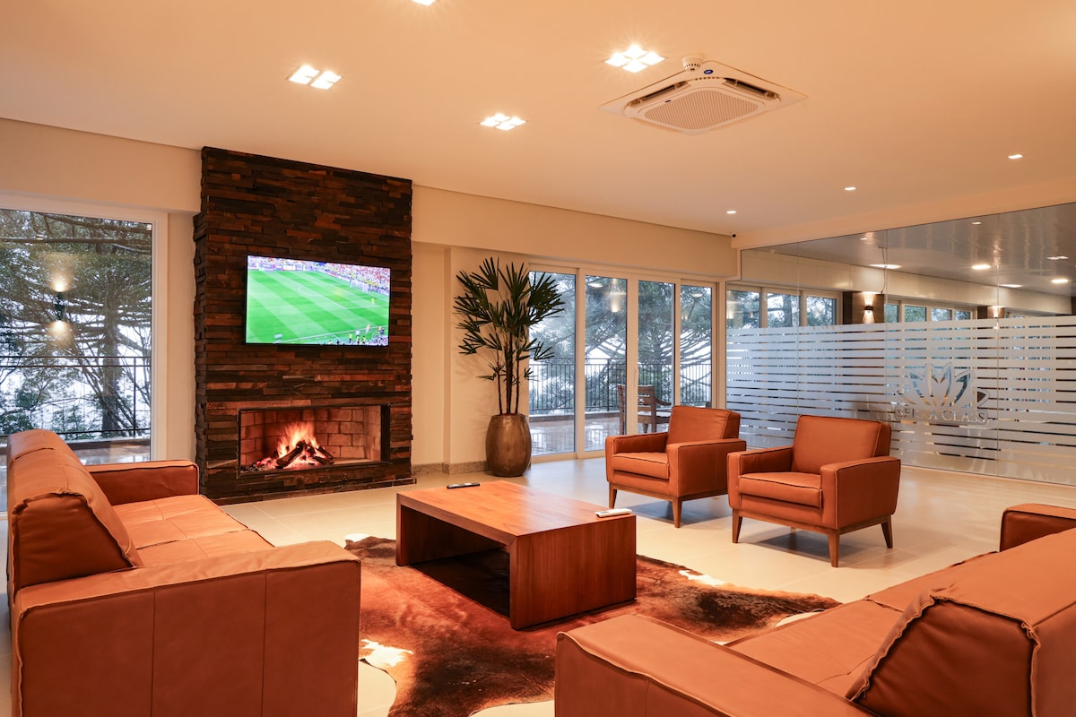 A spacious lounge area features a warm fireplace surrounded by a sofa and two armchairs. A large wooden coffee table is centrally placed. A television is mounted on the wall, and natural light streams through large windows, enhancing the modern design.
