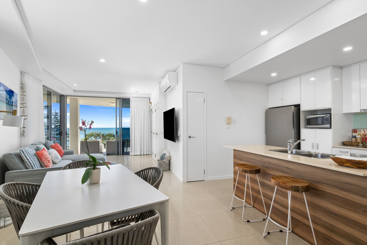 An open-concept living area is visible, featuring a modern kitchen with sleek cabinetry and a large island with bar stools. A dining table with woven chairs is nearby. Light fills the space, highlighting ocean views through expansive glass doors leading to a balcony.