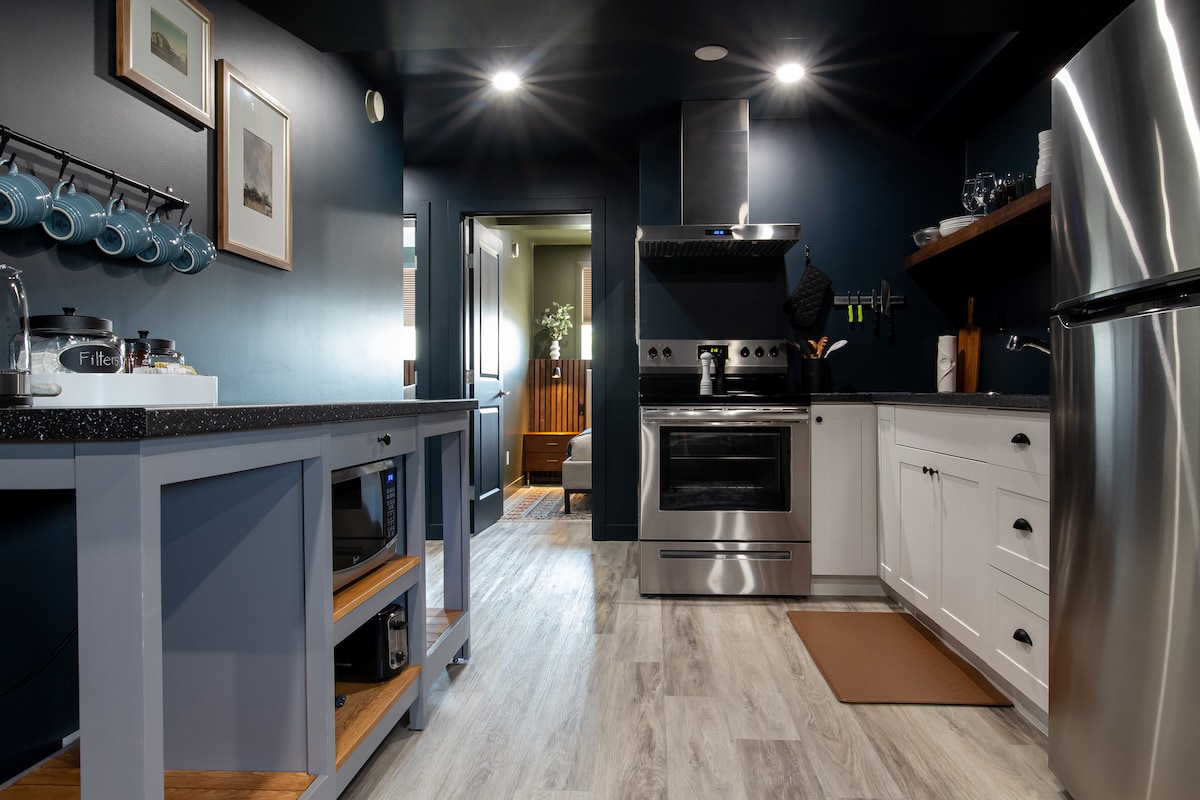A modern kitchen features dark blue walls and bright overhead lighting. Stainless steel appliances, including an oven and a microwave, are complemented by white cabinetry and an open shelving unit. Counter space is maximized with wooden accents, providing a functional and inviting cooking area.