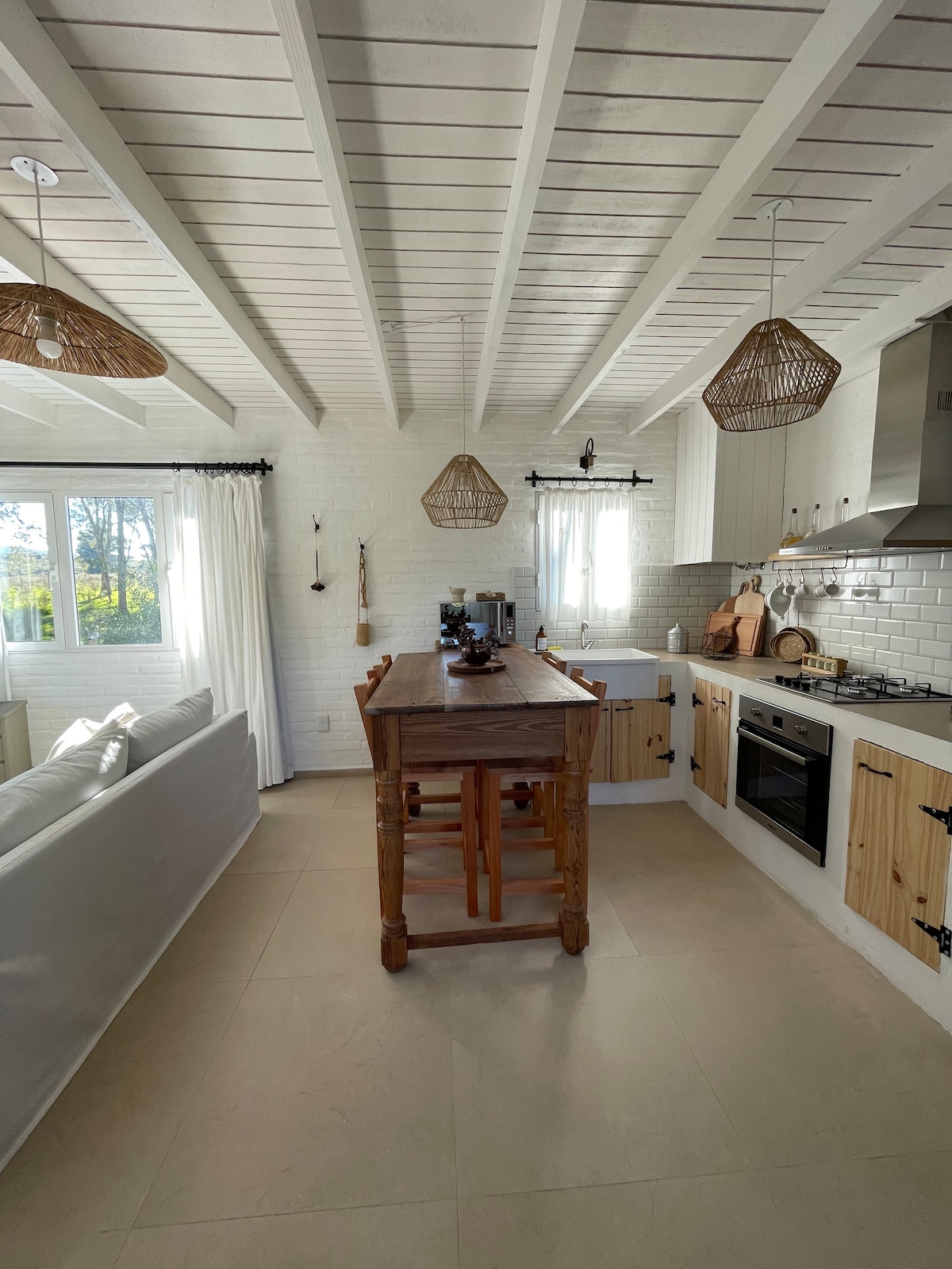 A bright kitchen area features a wooden island with seating, complemented by a sleek stove and modern appliances. White cabinetry and exposed beams add to the open feel. Natural light enters through large windows, framed by light curtains that provide a soft ambiance.