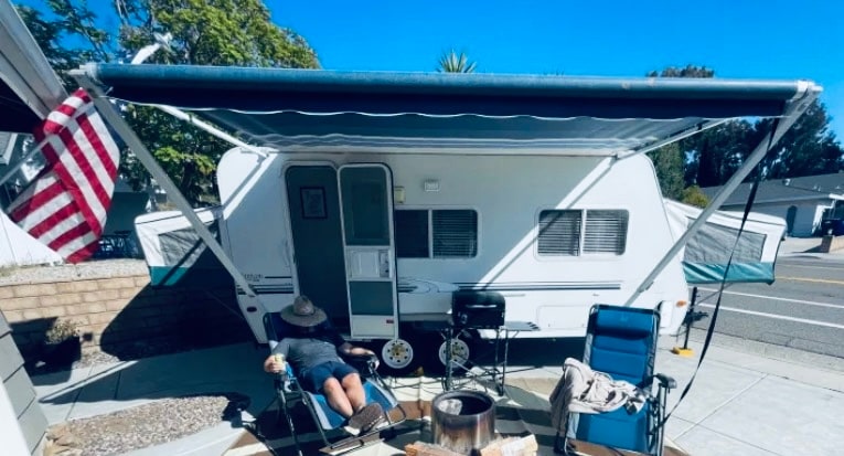 A 23-foot white camper is parked under a blue awning, with an American flag displayed. Two blue folding chairs and a fire pit surround it, providing a cozy outdoor gathering space. The background features a clear blue sky and a neighborhood setting.