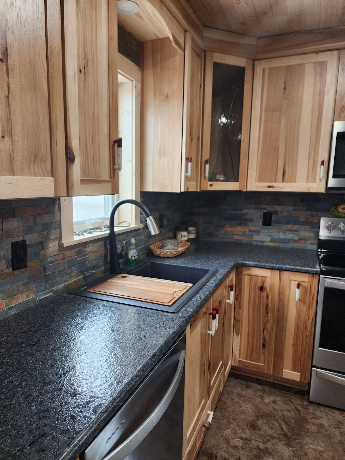 A modern kitchen area is showcased with natural wood cabinetry and a sleek granite countertop. A stainless steel sink is positioned beside a wooden cutting board, complemented by a textured stone backsplash. Soft lighting illuminates the space, enhancing the natural materials.