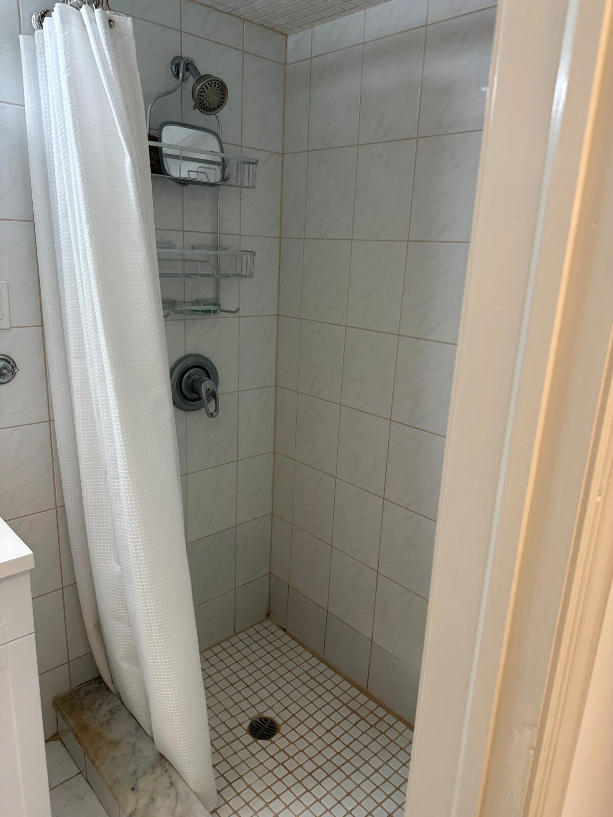 A clean shower space features tiled walls and a glass shelf for toiletries. The white curtain is drawn, providing privacy. A chrome showerhead is mounted above, and a small marble ledge is visible at the base of the shower.