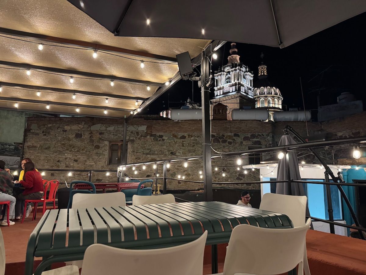A rooftop terrace is illuminated by string lights, creating a warm ambiance. Metal tables and chairs are arranged for seating, with a view of historical architecture in the background, including a church adorned with illuminated details, visible against the night sky.