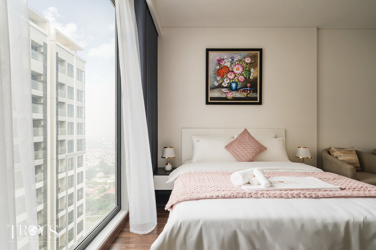A bright bedroom features a spacious bed adorned with soft linens and a pink decorative pillow. Floral artwork is mounted above the bed. Natural light enters through large windows, offering views of a nearby high-rise building. A small side table with lamps is positioned on either side of the bed.