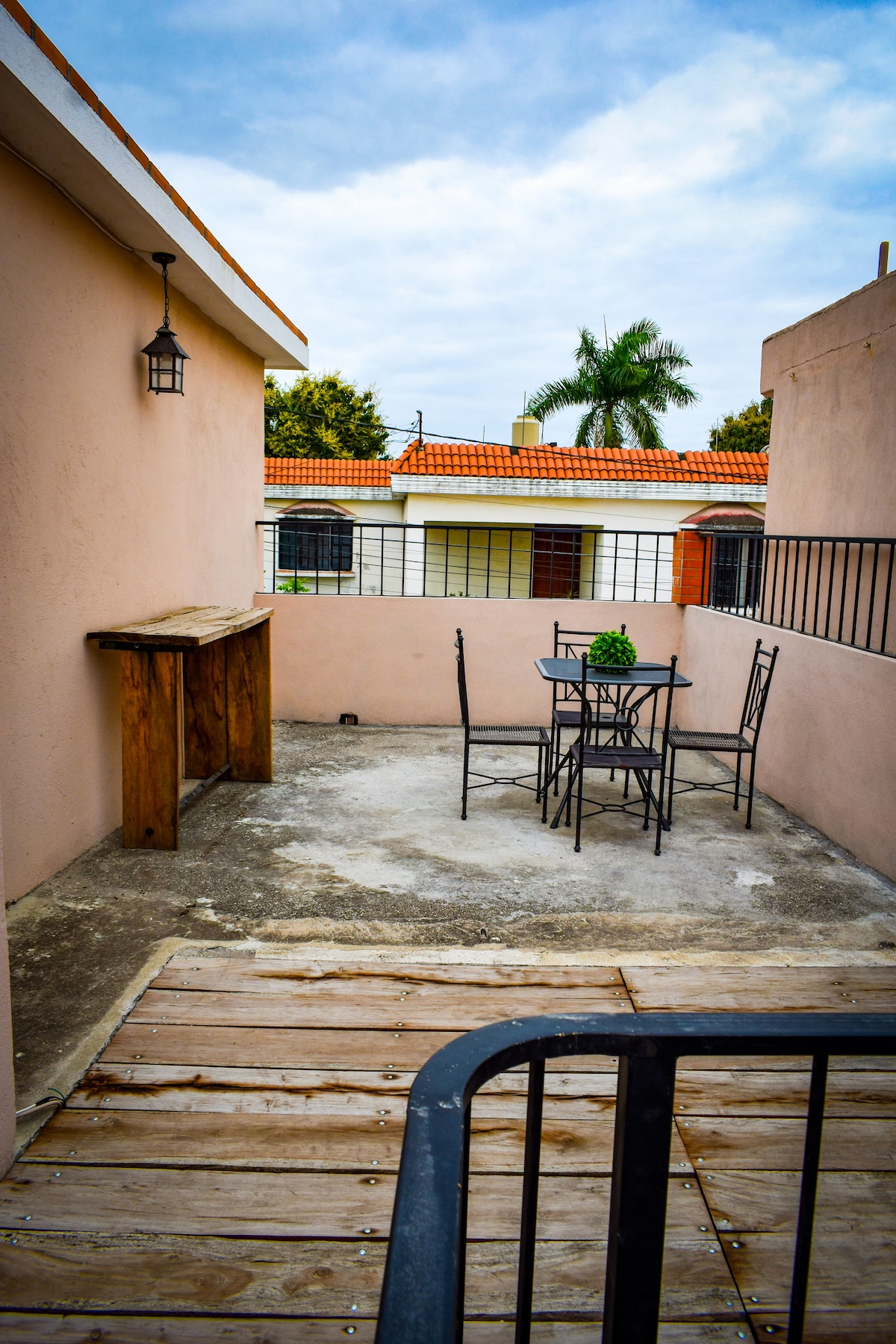 A small outdoor terrace features a wooden bar against one wall and a round metal table surrounded by four black chairs. The surface is a mix of concrete and wooden planks. A garden view is visible beyond the railing.