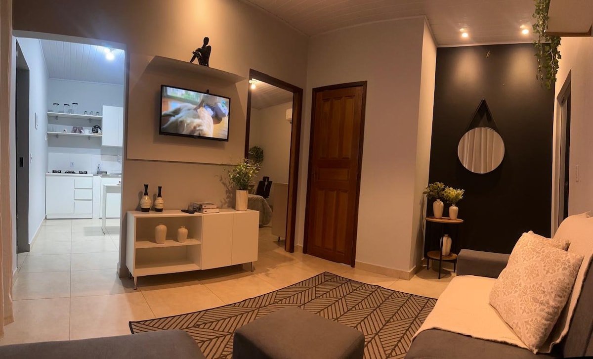 A cozy living area features a grey sofa adorned with decorative cushions. A wall-mounted television is positioned across from a white cabinet showcasing decorative items. Soft lighting complements the modern design, while a mirror adds depth to the space.