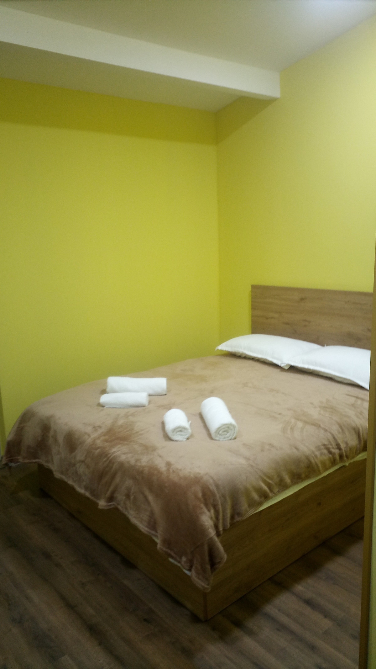 A cozy bedroom features a wooden bed with a soft beige blanket and white pillows. Freshly rolled towels are neatly placed on the bed. The walls are painted a bright yellow, creating a cheerful and inviting space.