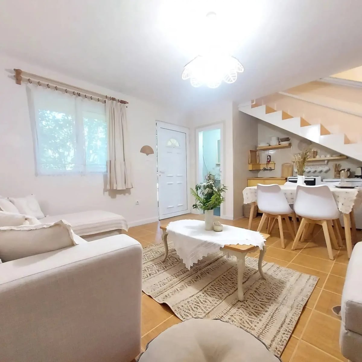 A welcoming living space features neutral-colored sofas arranged around a decorative coffee table. Natural light enters through window treatments, illuminating woven rugs and light-colored walls. A set of stairs leads to an upper level, with an open dining area visible in the background.