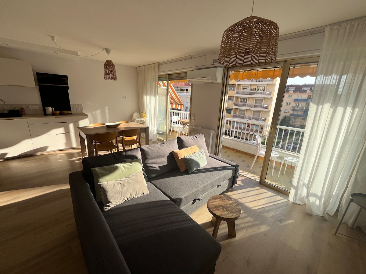 A bright living space is highlighted by large glass doors leading to a balcony. Sunlight fills the room, illuminating the modern dining area with a wooden table and chairs. A gray sofa is positioned comfortably, complemented by soft cushions and a small wooden side table.