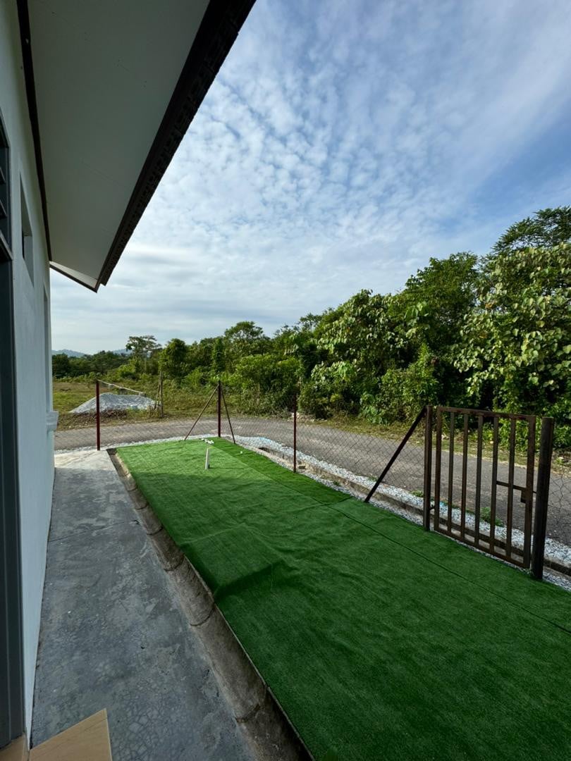 An outdoor area is presented, featuring a green artificial grass surface alongside a gravel pathway. A fence encloses the space, with trees and greenery visible in the background under a partially cloudy sky.