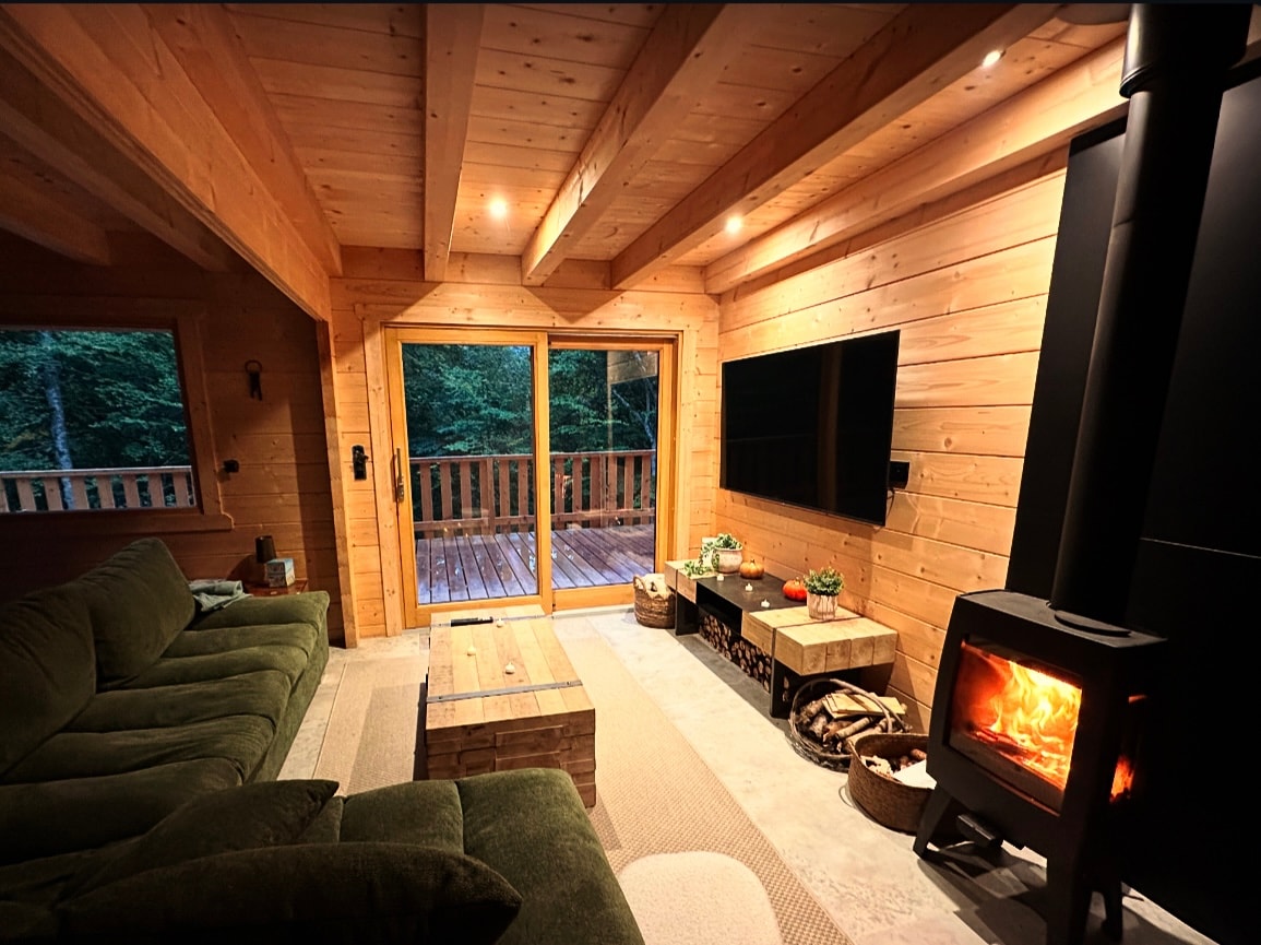 A cozy living area features a plush green sofa and a rustic coffee table. A television is mounted on the wooden wall, while a wood-burning stove provides warmth. Large sliding doors open to a deck, allowing natural light to fill the space.
