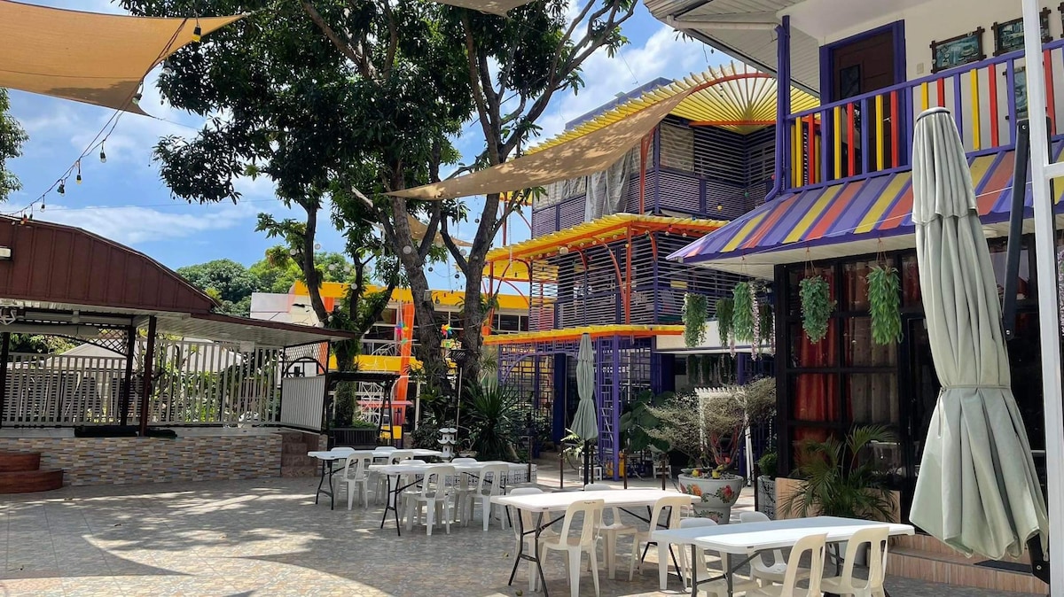 An outdoor area is framed by tall trees, providing shade over white tables and chairs arranged in a social setting. Colorful structures in the background feature vibrant shades of yellow and purple. Soft lighting adorns the space, contributing to a relaxed environment.