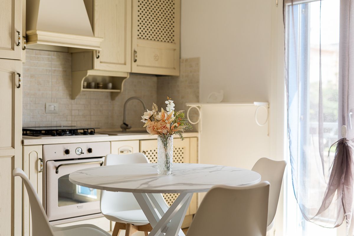 A light-filled kitchen area features a round dining table surrounded by four white chairs. A bouquet of flowers is placed in a vase on the table. The kitchen includes a gas stove and a well-organized countertop, providing a welcoming atmosphere for dining.