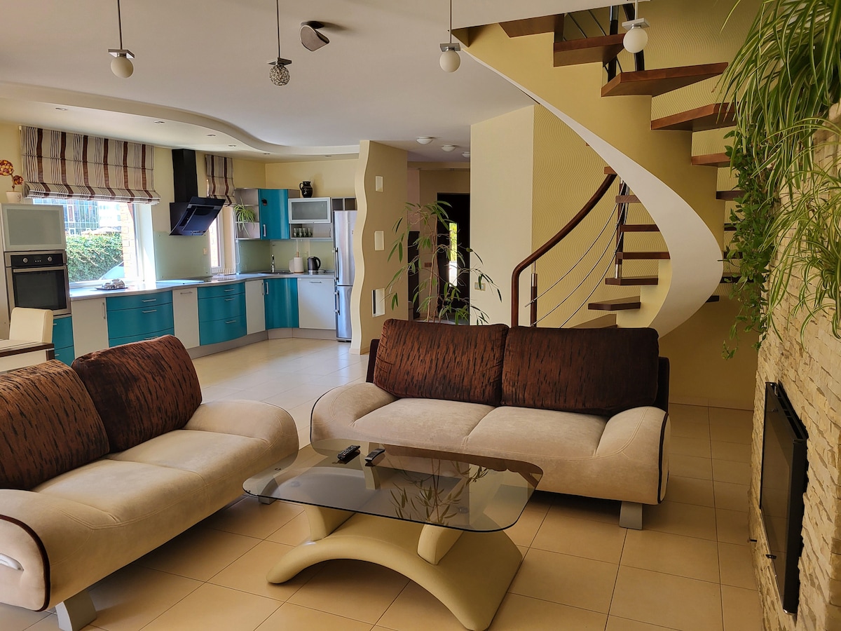 A spacious living area features two soft sofas and a glass coffee table, complemented by a modern staircase leading to the upper floor. A large window allows natural light to illuminate the room, and a sleek kitchen area with blue cabinetry is visible in the background.