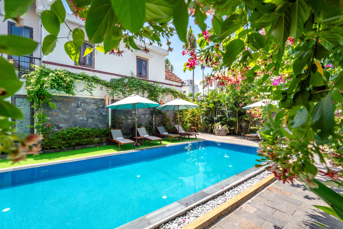 A large swimming pool is surrounded by vibrant greenery and tropical flowers. Lounge chairs are positioned under umbrellas along one side, providing shade and relaxation. The pool area is framed by natural stone walls, enhancing the serene atmosphere.