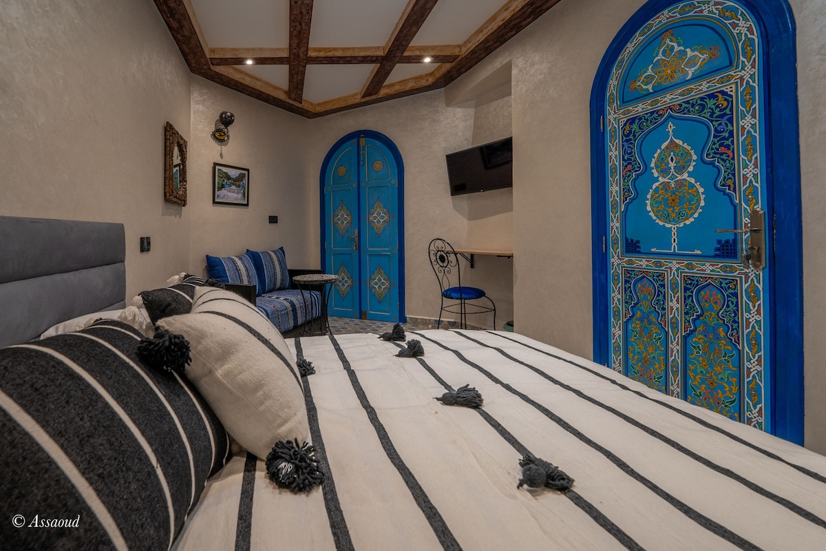 A comfortably arranged bedroom features a spacious bed adorned with striped linens and decorative cushions. Vibrant blue doors with intricate designs add character, while a flat-screen TV is mounted on the wall. A small seating area with a chair and rug completes the inviting space.