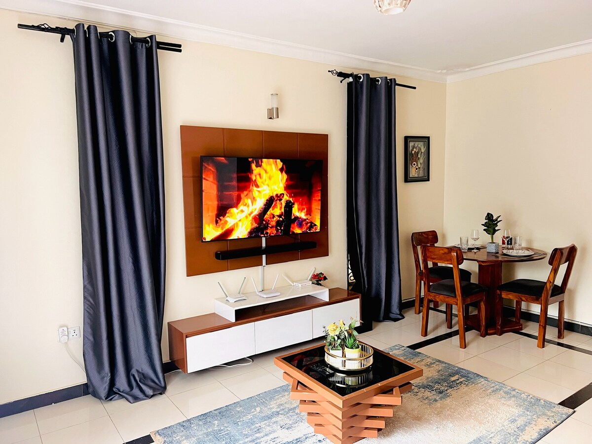 A living area features a modern wall-mounted television displaying a fireplace effect. Dark curtains frame the window, and a stylish coffee table occupies the center. A dining set with wooden chairs is positioned against the wall, complemented by subtle decorative accents.